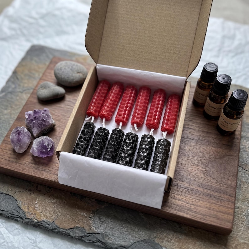 May include: A cardboard box containing twelve cylindrical beeswax candles with a honeycomb texture, arranged in two rows of six, with red candles on top and black candles on the bottom. Three small essential oil bottles and three amethyst crystals are nearby.