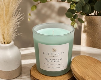 May include: A light green soy candle in a frosted glass jar with a white label reading LIFE ARIS PEPERMINT AND EUCALYPTUS HANDCRAFTED SOY CANDLE Made in Missouri, USA. The candle sits on a round wooden coaster next to a circular bamboo lid.