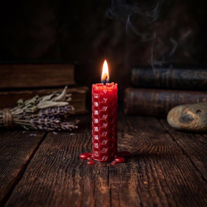 May include: A cylindrical red beeswax candle with a textured honeycomb pattern. The candle is lit, featuring a bright flame and a thin trail of smoke rising from the wick. Melted red wax has pooled around the base of the candle.