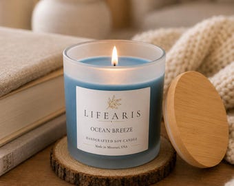 May include: A frosted glass candle with blue wax and a lit wick, featuring a white label that reads LIFEARIS OCEAN BREEZE HANDCRAFTED SOY CANDLE Made in Missouri, USA. The candle sits on a circular wood slice coaster next to a round wooden lid.