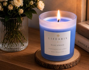 May include: A blue soy candle in a frosted glass jar with a lit wick, featuring a white label that reads LIFEARIS BLUE SPRUCE HANDCRAFTED SOY CANDLE Made in Missouri, USA. The candle sits on a circular wooden coaster with visible bark edges.