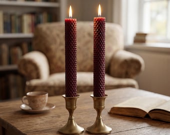 May include: Two tall, dark red, cylindrical beeswax candles with a textured honeycomb pattern, each lit with a steady flame. The candles are held upright in matching, polished brass candlestick holders with a flared base and a slender stem.