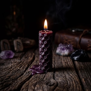 May include: A dark purple pillar candle with a textured honeycomb pattern. The candle is lit, with a bright flame at the top and a small amount of melted wax pooling at the base. The wax has a deep, rich color and a distinct hexagonal surface design.