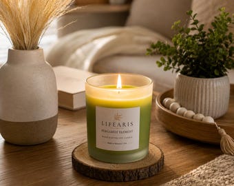 May include: A lit soy candle in a frosted green glass jar with a white label reading LIFEARIS BERGAMOT ELEMENT HANDCRAFTED SOY CANDLE Made in Missouri, USA. The candle sits on a circular wood slice coaster with a visible bark edge.