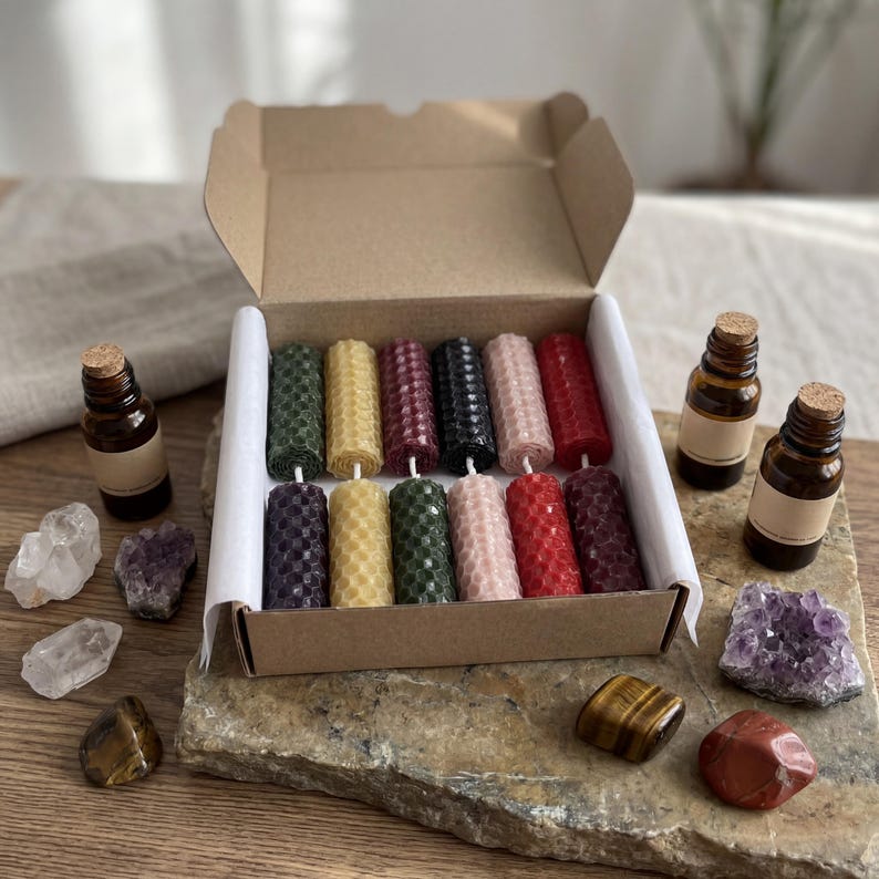 May include: A cardboard box containing twelve cylindrical beeswax candles with a honeycomb texture in various colors, including green, yellow, red, black, and pink. Two amber glass bottles with cork stoppers and several raw crystals are arranged nearby.