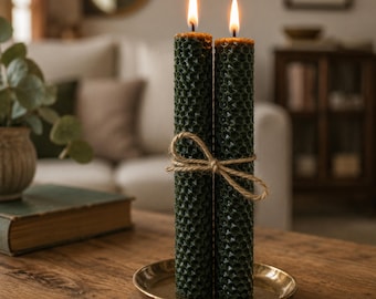 May include: Two dark green beeswax candles with a honeycomb pattern, tied together with twine, are lit and placed on a small, round, gold-colored tray. The candles are on a wooden table, with a blurred background of a living room.