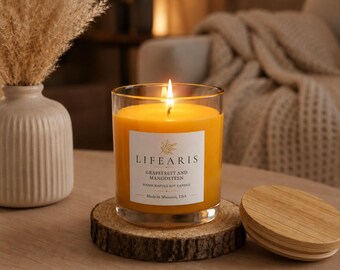 May include: A clear glass jar candle with bright orange wax and a lit wick. The white label reads LIFEARIS, GRAPEFRUIT AND MANGOSTEEN, HANDCRAFTED SOY CANDLE, Made in Missouri, USA. The candle sits on a round wood slice coaster with a matching lid nearby.