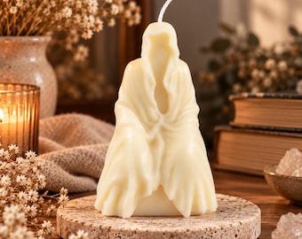May include: A cream-colored wax candle shaped like a hooded figure draped in a flowing robe. The candle features a single white wick extending from the top of the hood. It is positioned on a circular, textured, light brown stone-like base.