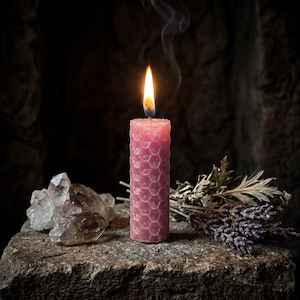 May include: A pink cylindrical candle with a textured honeycomb pattern is lit, emitting a small flame and a thin trail of smoke. Beside the candle are clear quartz crystal clusters and a bundle of dried lavender sprigs.