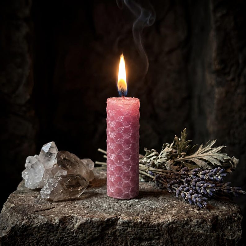 May include: A pink cylindrical candle with a textured honeycomb pattern is lit, emitting a small flame and a thin trail of smoke. Beside the candle are clear quartz crystal clusters and a bundle of dried lavender sprigs.