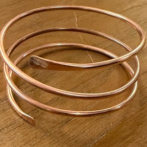 May include: A copper-colored spiral bracelet. The bracelet wraps around the wrist multiple times, with a simple, elegant design. The metal has a warm, reddish-brown hue, and the ends are slightly flattened.