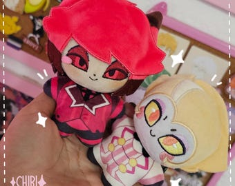 Puede incluir: Dos peluches con personajes estilizados. Uno tiene el pelo rojo brillante, ojos rojos y un conjunto rojo y negro. El otro tiene el pelo amarillo claro, ojos amarillos y un conjunto blanco con detalles circulares en rosa y amarillo.
