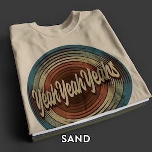 May include: A sand-colored t-shirt featuring a circular graphic with concentric rings in shades of blue, tan, and rust. The text "Yeah Yeah Yeahs" is printed in a cursive, dark brown font across the center of the design, which has a distressed, weathered texture.