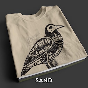 Può includere: Una t-shirt color sabbia con una grafica nera di un uccello. Il corpo dell'uccello contiene il testo: I don't mind stealing bread from the mouths of decadence. Il design ha un aspetto usurato e testurizzato stampato sulla parte anteriore.