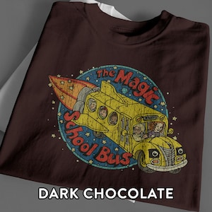 Op de afbeelding: Een donker chocoladebruin t-shirt met een afbeelding van een gele schoolbus in de vorm van een raket. Het ontwerp bevat de tekst The Magic School Bus in het rood, omringd door een blauwe cirkelvormige achtergrond met sterren en planeten.