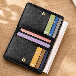May include: Black bi-fold wallet with multiple card slots, filled with colorful credit cards. The wallet is open, revealing its interior. A cup of coffee and a notebook with the word "NOTES" are also visible.