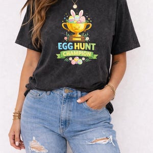 Op de afbeelding: Donkergrijs T-shirt met een gouden trofee, konijnenoren en kleurrijke eieren, met de tekst "EGG HUNT CHAMPION" op een groene banner. Het shirt wordt gedragen met lichtblauwe ripped jeans.