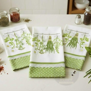 Pode incluir: Três panos de cozinha brancos com ervas e flores verdes bordadas. As toalhas têm uma borda de bolinhas verdes com uma borda de renda branca. Potes de especiarias ao fundo.