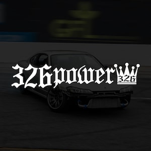May include: A dark grey sports car with the text "326power" in a gothic font. The car has a modified front end and blue accents. The background is blurred, suggesting motion.