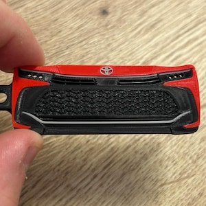 Może przedstawiać: Czerwono-czarny plastikowy brelok w kształcie przedniego grilla pojazdu, z wyraźnym czarnym wzorem plastra miodu, srebrnym logo Toyoty na środku u góry oraz okrągłym otworem do mocowania po lewej stronie.