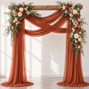 Pode incluir: Um arco de casamento de madeira drapeado com tecido transparente cor de terracota, apresentando dois grandes arranjos florais nos cantos superiores. Os buquês consistem em rosas creme, bege e laranja queimado, acentuadas com folhas de eucalipto.