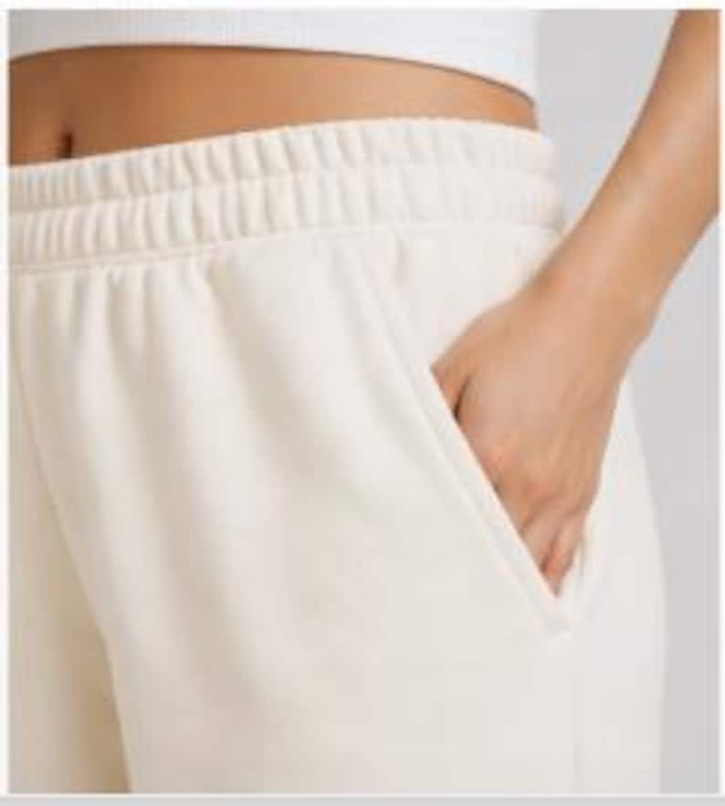 Puede incluir: Pantalones de ch&aacute;ndal color crema con cintura el&aacute;stica y bolsillos. La imagen muestra un primer plano de la cintura y la zona de los bolsillos, con una mano dentro de uno de los bolsillos. La tela parece suave y c&oacute;moda.