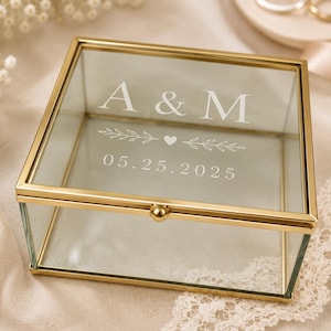 May include: A square glass keepsake box with a gold metal frame. The lid features white etched text reading A & M, a small heart icon flanked by leafy branches, and the date 05.25.2025. The box has a small gold knob on the front edge.