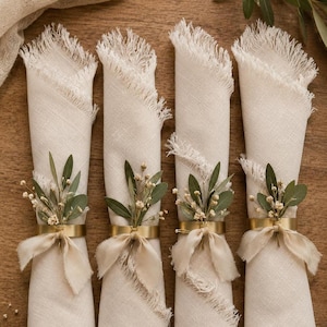 May include: Four rolled cream linen napkins with fringed edges, each decorated with a gold ring, a beige ribbon, and sprigs of greenery. The napkins are arranged on a wooden surface, ideal for a table setting.