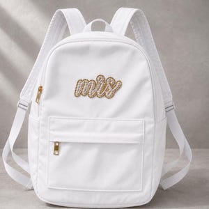 Puede incluir: Una mochila blanca con cremallera dorada y un parche central con la palabra "mrs" escrita en cursiva, adornada con pequeñas perlas blancas. La mochila incluye correas ajustables para los hombros y un bolsillo frontal con cremallera.