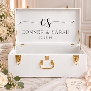 May include: A white wooden keepsake box with gold-toned metal latches and a handle. The interior lid features black printed text that reads CS, CONNER & SARAH, and 12.26.26. The box is open, revealing a spacious rectangular storage compartment.