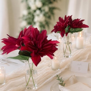 Può includere: Un'apparecchiatura con fiori bordeaux in vasi di vetro trasparente. Il tavolo è decorato con candele, un runner color crema e un segnaposto con il numero del tavolo. I fiori sono il punto focale, aggiungendo un tocco di eleganza.