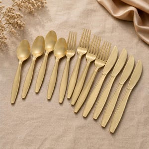 May include: A collection of gold-colored flatware, including spoons, forks, and knives, arranged on a neutral-toned surface. The utensils have a matte finish and a simple, elegant design, suitable for formal dining or special occasions.