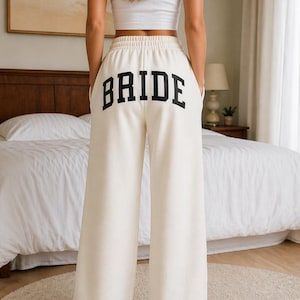Puede incluir: Pantalones deportivos color crema de pierna ancha con la palabra "BRIDE" impresa en letras negras en negrita en la parte trasera. Los pantalones tienen cintura con cordón y bolsillos. La modelo lleva una camiseta sin mangas blanca.