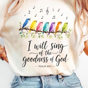 May include: An ivory t-shirt featuring a graphic of seven colorful birds perched on a branch with musical notes above them. The text reads, I will sing of the goodness of God, followed by PSALM 89:1 in a black script and serif font combination.