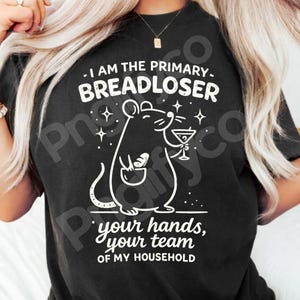 Könnte beinhalten: Ein schwarzes Kurzarm-T-Shirt mit einer weißen Grafik einer Maus, die einen Laib Brot und ein Martiniglas hält. Der Text lautet: I AM THE PRIMARY BREADLOSER your hands, your team OF MY HOUSEHOLD. Das Design enthält kleine Sternakzente.