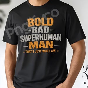 Può includere: T-shirt nero con la scritta "BOLD BAD SUPERHUMAN MAN - THAT'S JUST WHO I AM!" in arancione e grigio. La maglietta è a girocollo.