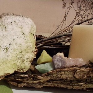 Puede incluir: Una exhibici&oacute;n r&uacute;stica y natural con una piedra grande y texturizada con acentos verdes, una vela color crema y cristales coloridos. El arreglo est&aacute; sobre un trozo de corteza, con ramas secas y vegetaci&oacute;n.