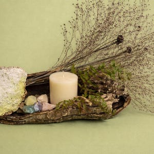 Puede incluir: Arreglo decorativo r&uacute;stico con una vela gruesa de color crema, peque&ntilde;os cristales coloridos y plantas secas. Los elementos est&aacute;n dispuestos en un trozo de corteza, con una piedra grande y texturizada a la izquierda. Fondo verde claro.