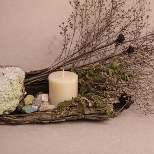 Puede incluir: Una exhibici&oacute;n r&uacute;stica con una vela de pilar crema, gemas coloridas y ramas secas. El arreglo est&aacute; sobre un trozo de corteza texturizada, creando una est&eacute;tica natural y terrosa. El fondo es de un tono suave y neutro.