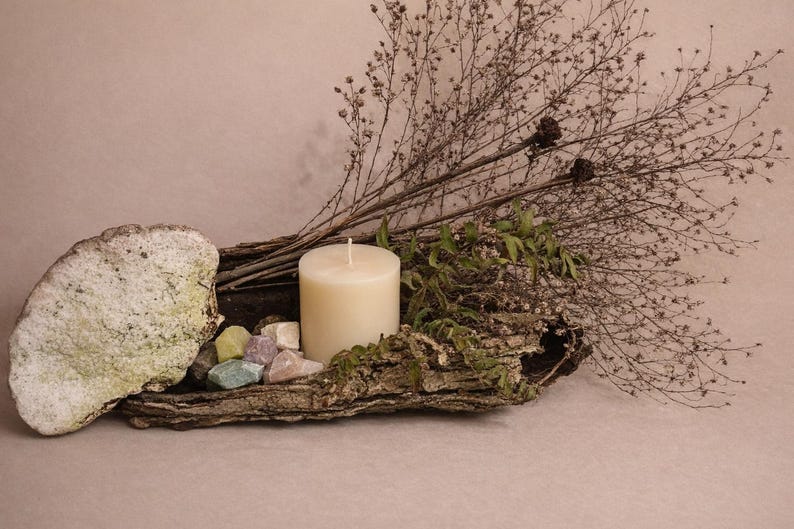 Puede incluir: Una exhibici&oacute;n r&uacute;stica con una vela de pilar crema, gemas coloridas y ramas secas. El arreglo est&aacute; sobre un trozo de corteza texturizada, creando una est&eacute;tica natural y terrosa. El fondo es de un tono suave y neutro.