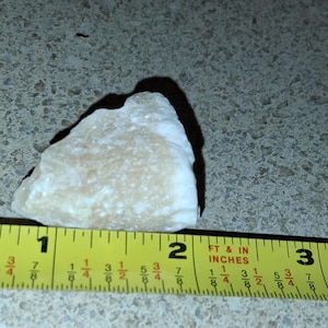 Puede incluir: Una roca de color claro, de forma irregular, de aproximadamente 5 cm de largo, se mide con una cinta m&eacute;trica amarilla. La roca tiene una textura rugosa y proyecta una sombra. La cinta m&eacute;trica est&aacute; marcada en pulgadas y fracciones.