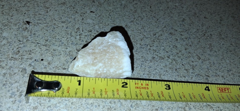 Puede incluir: Una roca de color claro, de forma irregular, de aproximadamente 5 cm de largo, se mide con una cinta m&eacute;trica amarilla. La roca tiene una textura rugosa y proyecta una sombra. La cinta m&eacute;trica est&aacute; marcada en pulgadas y fracciones.