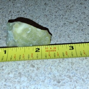 Puede incluir: Una piedra verde claro de forma irregular se encuentra junto a una cinta m&eacute;trica amarilla. La piedra mide aproximadamente 5 cm de largo. La cinta m&eacute;trica muestra medidas en pulgadas y pies.