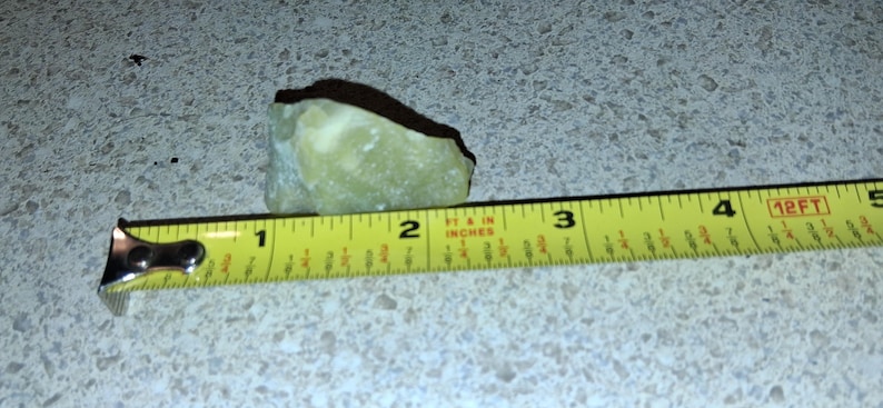 Puede incluir: Una piedra verde claro de forma irregular se encuentra junto a una cinta m&eacute;trica amarilla. La piedra mide aproximadamente 5 cm de largo. La cinta m&eacute;trica muestra medidas en pulgadas y pies.
