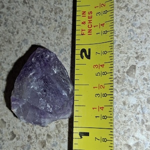 Puede incluir: Un cristal de amatista p&uacute;rpura en bruto junto a una cinta m&eacute;trica amarilla. El cristal mide aproximadamente 5 cm de largo. La cinta m&eacute;trica muestra pulgadas y fracciones de pulgada.