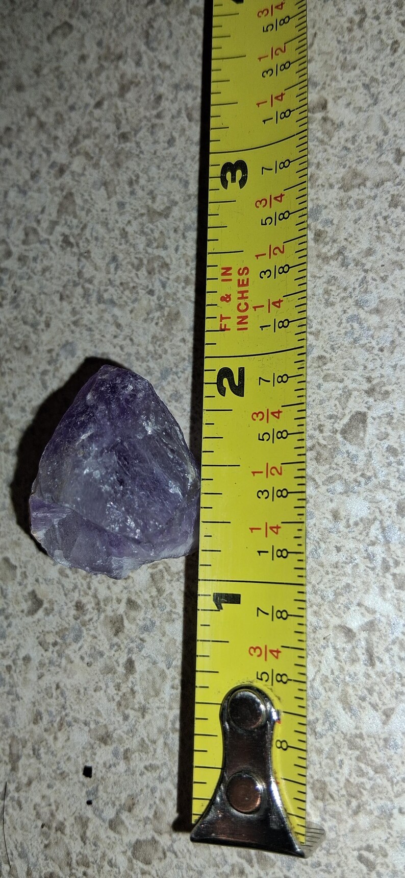 Puede incluir: Un cristal de amatista p&uacute;rpura en bruto junto a una cinta m&eacute;trica amarilla. El cristal mide aproximadamente 5 cm de largo. La cinta m&eacute;trica muestra pulgadas y fracciones de pulgada.