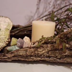 Puede incluir: Arreglo decorativo r&uacute;stico con una vela color crema, piedras preciosas coloridas y follaje seco. La vela se asienta sobre una base cubierta de corteza, con una piedra grande y texturizada a la izquierda. Una est&eacute;tica natural y terrosa.
