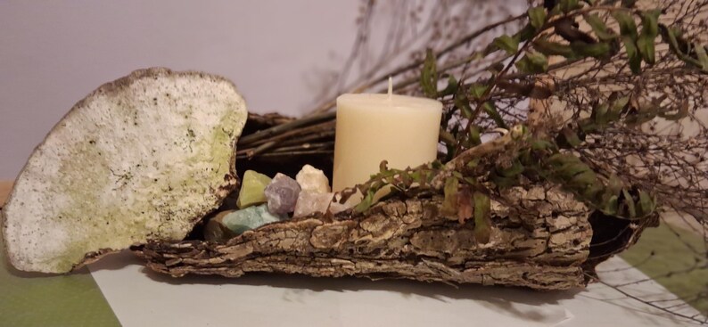 Puede incluir: Arreglo decorativo r&uacute;stico con una vela color crema, piedras preciosas coloridas y follaje seco. La vela se asienta sobre una base cubierta de corteza, con una piedra grande y texturizada a la izquierda. Una est&eacute;tica natural y terrosa.