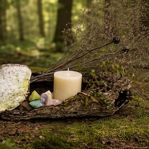 Puede incluir: Arreglo natural con una vela de pilar crema, cristales coloridos y follaje seco en un recipiente de madera r&uacute;stico. Una piedra grande y texturizada se encuentra a la izquierda, todo sobre un fondo de bosque borroso.