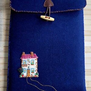 May include: A navy blue felt laptop sleeve with a stitched house design. The sleeve has a brown leather triangle closure with a wooden toggle. The edges are stitched with a contrasting brown thread.